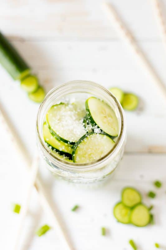 Sliced cucumbers with salt in a large mason jar on a white counter.