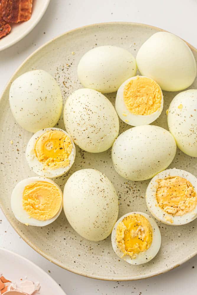 Hard boiled eggs, some whole, some cut in half, sprinkled with pepper and on a white plate. Side plate with some bacon on it.