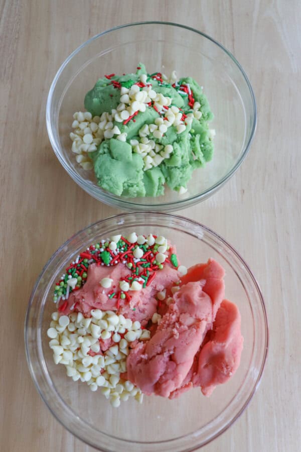 Pink and green cookie dough covered in holiday sprinkles and white chocolate chips in two separate bowls.