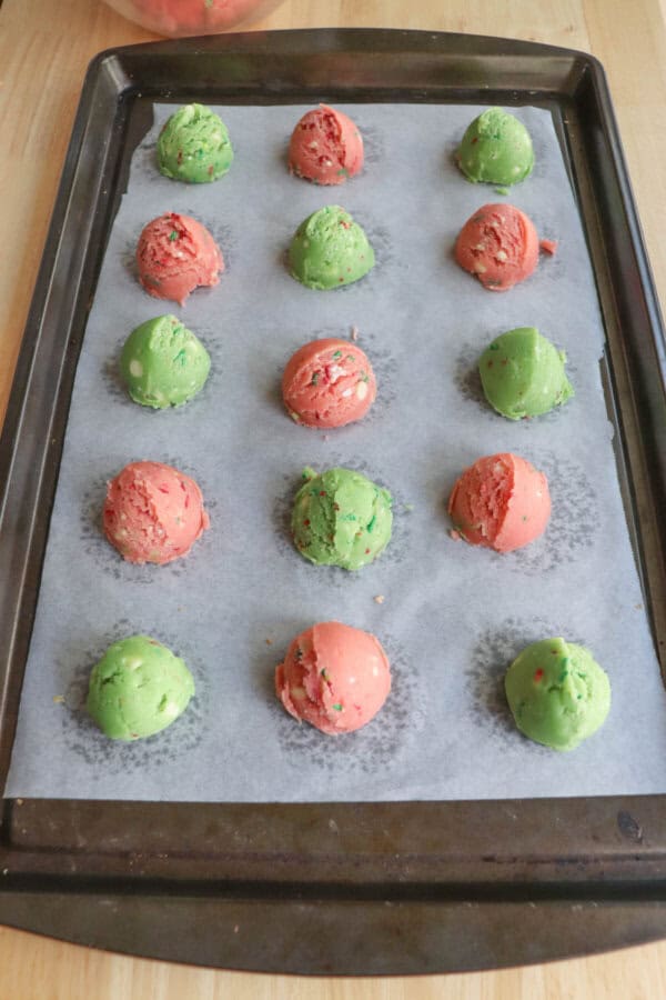 Pink and green balls of cookie dough on a parchment paper lined baking sheet.