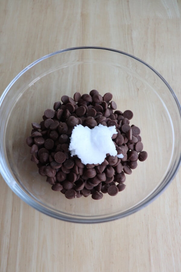 Chocolate chips and coconut oil in a small clear glass bowl on a wooden table.