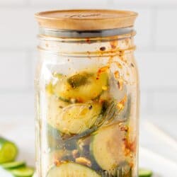 Sliced cucumbers, chili dressing and sesame seeds in a clear glass jar on a white counter with a white tile background.