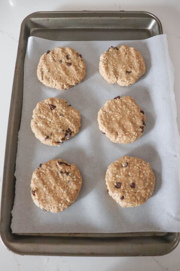 Six large rounds of oatmeal chocolate chip cookie dough on a parchment paper lined baking sheet.