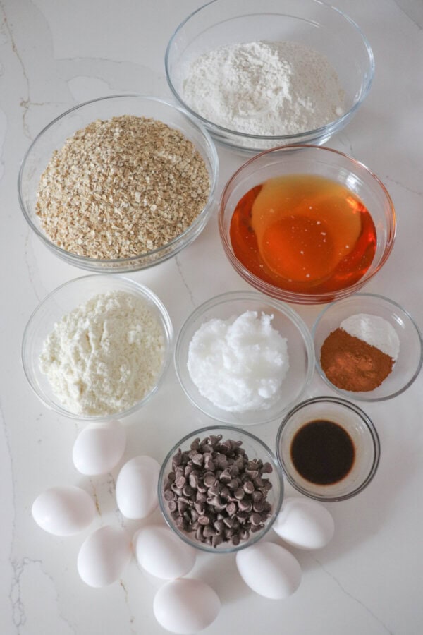 Ingredients for oatmeal protein cookies in clear glass bowls on a white ocunter.