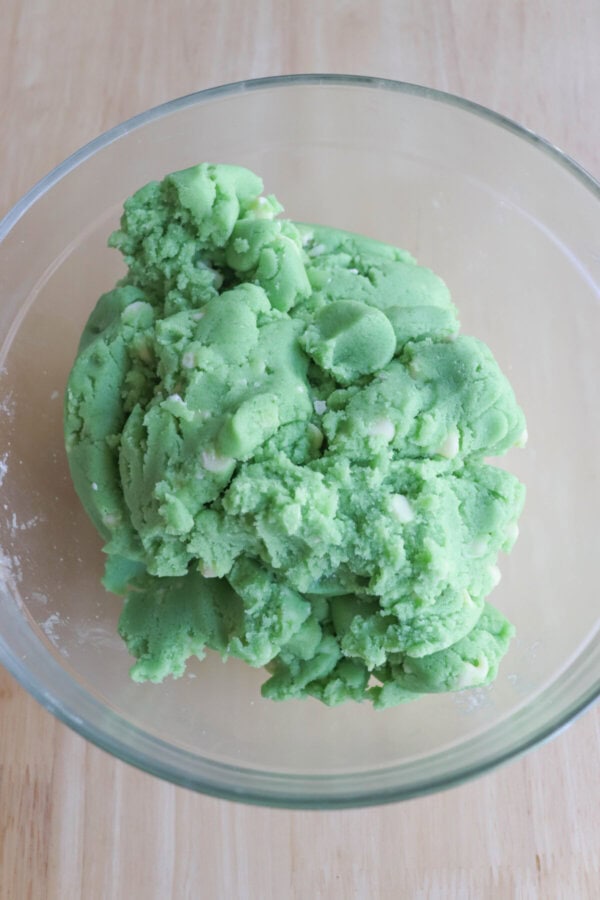 Green cookie dough in a clear glass bowl on a wooden table.