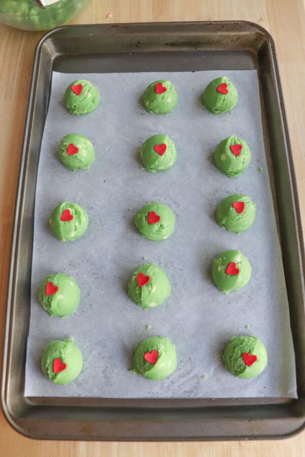 Green balls of cookie dough with red candy hearts on a parchment paper lined cookie sheet.
