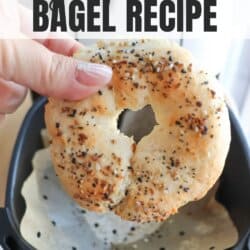 A hand holding a seasoned bagel above a parchment-lined air fryer basket. The text reads high protein bagel recipe. Below is the website name: homemadeheather.com.