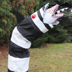 A large, striped black and white sculpture resembling a snake or worm stands upright on grassy ground. It has a jagged, open mouth with sharp red teeth and a horn, surrounded by tall trees.