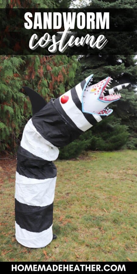 A large, striped black and white sculpture resembling a snake or worm stands upright on grassy ground. It has a jagged, open mouth with sharp red teeth and a horn, surrounded by tall trees.