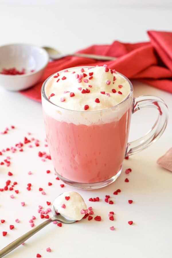 Red velvet hot chocolate in a clear mug with whipped cream and red sprinkles on top. There is a spoon with whipped cream and sprinkles, a small prep bowl with sprinkles and a red napkin nearby.
