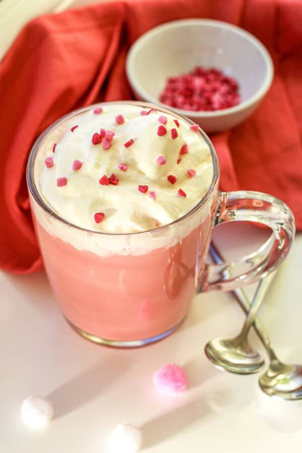 Red velvet hot chocolate in a clear mug with whipped cream and red sprinkles on top. There are two spoons, a small prep bowl with red & pink heart shaped sprinkles and a red napkin nearby.
