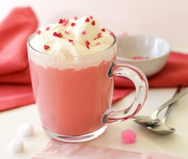 Red velvet hot chocolate in a clear mug with whipped cream and red sprinkles on top. There is are two spoons, a small prep bowl with sprinkles and a red napkin nearby.