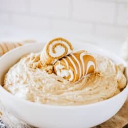 Creamy pumpkin spice dip in a white bowl topped with Little Debbie pumpkin rolls on a newspaper lined table.