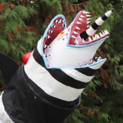 A large, striped black and white sculpture resembling a snake or worm stands upright on grassy ground. It has a jagged, open mouth with sharp red teeth and a horn, surrounded by tall trees.