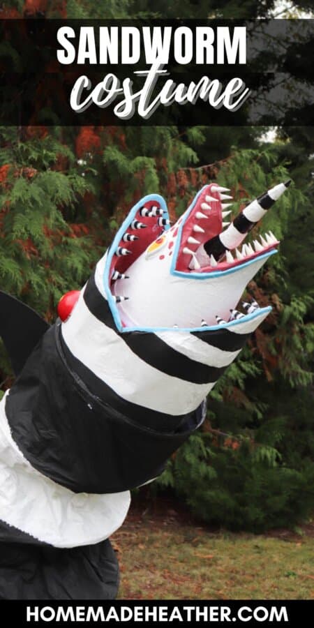 A large, striped black and white sculpture resembling a snake or worm stands upright on grassy ground. It has a jagged, open mouth with sharp red teeth and a horn, surrounded by tall trees.