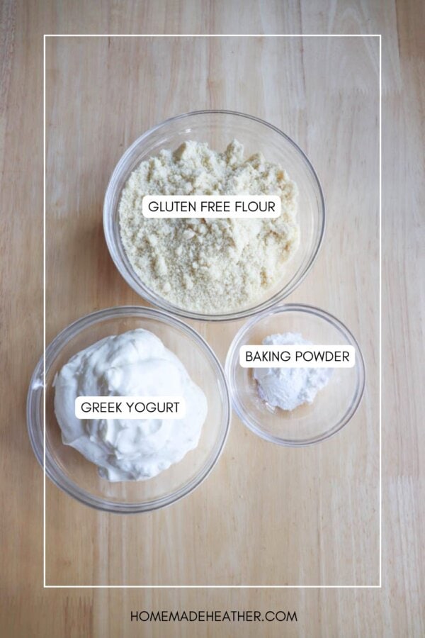 Three glass bowls on a wooden surface. The bowls contain labeled ingredients: Gluten Free Flour, Greek Yogurt, and Baking Powder. The website homemadeheather.com is visible at the bottom.
