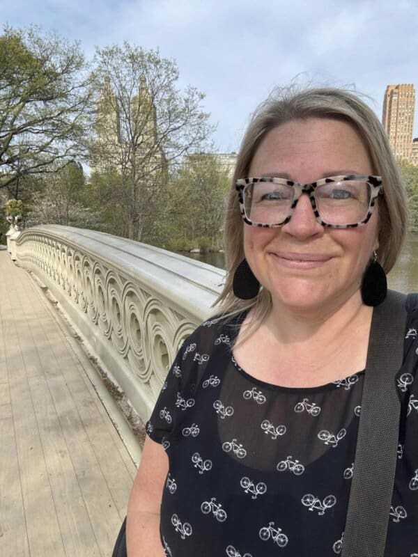 A woman with glasses, wearing a black shirt adorned with homemade bicycle patterns, stands gracefully on a decorative bridge. Trees and tall buildings form the backdrop on a sunny day, casting heather hues across the landscape.