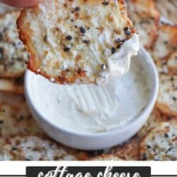 A hand holding a circular cottage cheese chip with seasoning, partially dipped in a bowl of white dip. More chips are scattered in the background. Text at the bottom reads cottage cheese chip recipe with a website link.