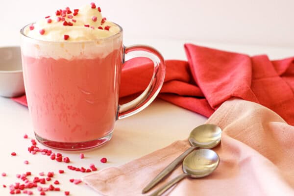 Red velvet hot chocolate in a clear mug with whipped cream and red sprinkles on top. There are two spoons, a small prep bowl with red & pink heart shaped sprinkles and red & pink napkins nearby.