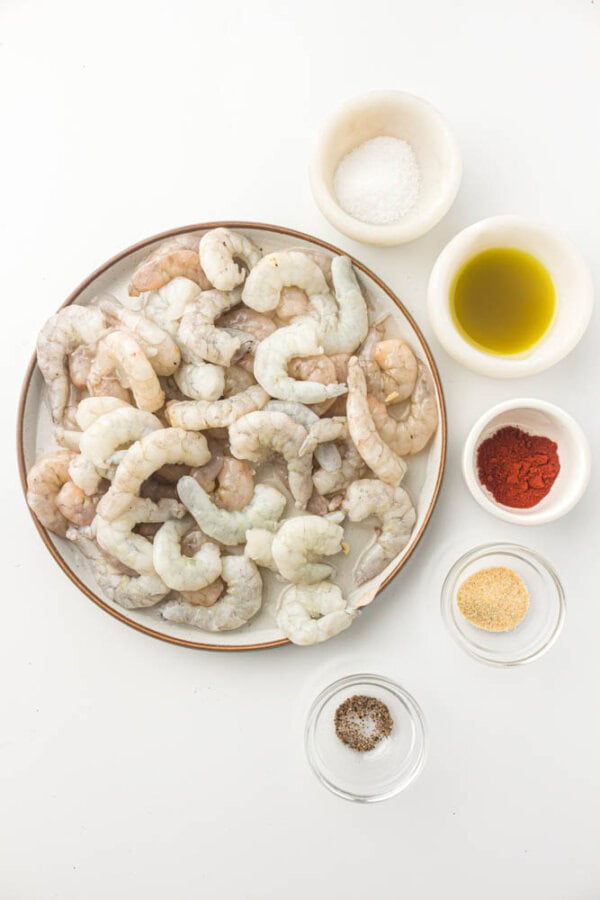 A plate of raw shrimp is surrounded by small bowls containing salt, olive oil, paprika, garlic powder, and pepper on a white background.