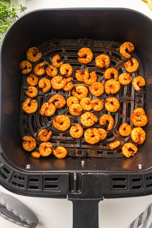 Cooked shrimp seasoned with spices are arranged in the basket of an air fryer. The shrimp appear golden and evenly cooked. The basket sits on a white surface, and fresh herbs are visible in the background.