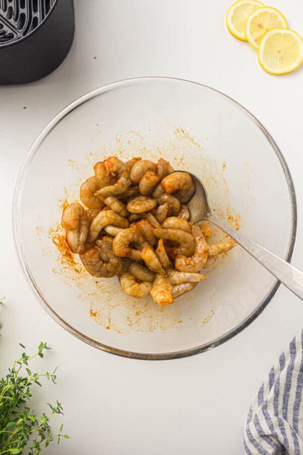 A glass bowl filled with seasoned shrimp, placed on a white surface. A silver spoon rests in the bowl. Slices of lemon, an air fryer, thyme, and a striped cloth are nearby.