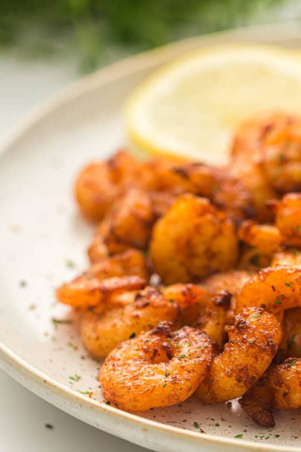 Close-up of a plate featuring grilled shrimp seasoned with spices, garnished with chopped herbs. A lemon wedge is placed in the background. The shrimp are golden-brown and appear appetizing.