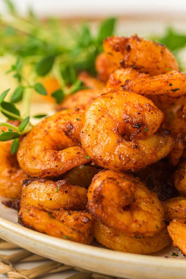Close-up of a plate of seasoned, grilled shrimp garnished with fresh herbs. The shrimp have a golden-brown, slightly charred appearance, indicating they are cooked with spices and herbs.