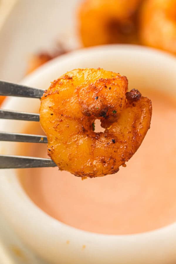 A fork holds a seasoned and grilled shrimp above a bowl of creamy, light orange dipping sauce. The shrimp is golden brown with visible spices, suggesting a flavorful preparation. Blurred background hints at additional food items.
