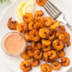 A plate of seasoned shrimp is displayed alongside a small bowl of sauce. Two lemon wedges and a fork are placed beside the shrimp. The dish is garnished with herbs, and everything is set on a white background.