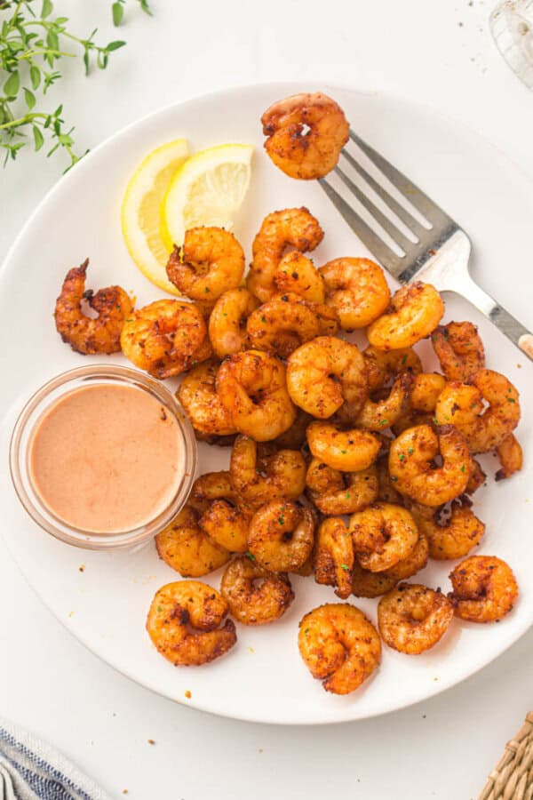 A plate of seasoned shrimp is displayed alongside a small bowl of sauce. Two lemon wedges and a fork are placed beside the shrimp. The dish is garnished with herbs, and everything is set on a white background.