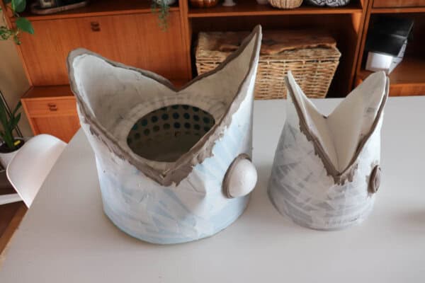 A white tabletop displaying two masks with large mouths covered in masking tape. A wooden basket and shelves are visible in the background.