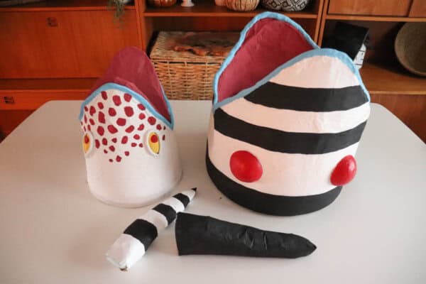Two handcrafted masks are displayed on a table. The left mask is white with red spots and yellow eyes, while the right mask features black and white stripes with red circles. Two matching striped accessories lie in front of the masks.
