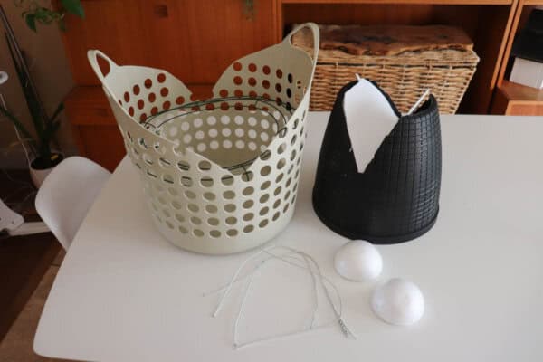 A white tabletop displaying a beige plastic basket, a black plastic container, two white foam balls on a circular wire frame, and two metal hangers. A wooden basket and shelves are visible in the background.