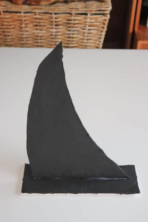 A simplistic black paper sculpture resembling a fin, standing vertically on a white surface. In the background, there are wicker baskets and wooden furniture.