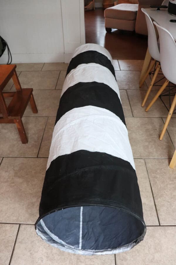A black and white striped play tunnel is laid out on a tiled floor in a room with chairs and a small table nearby.