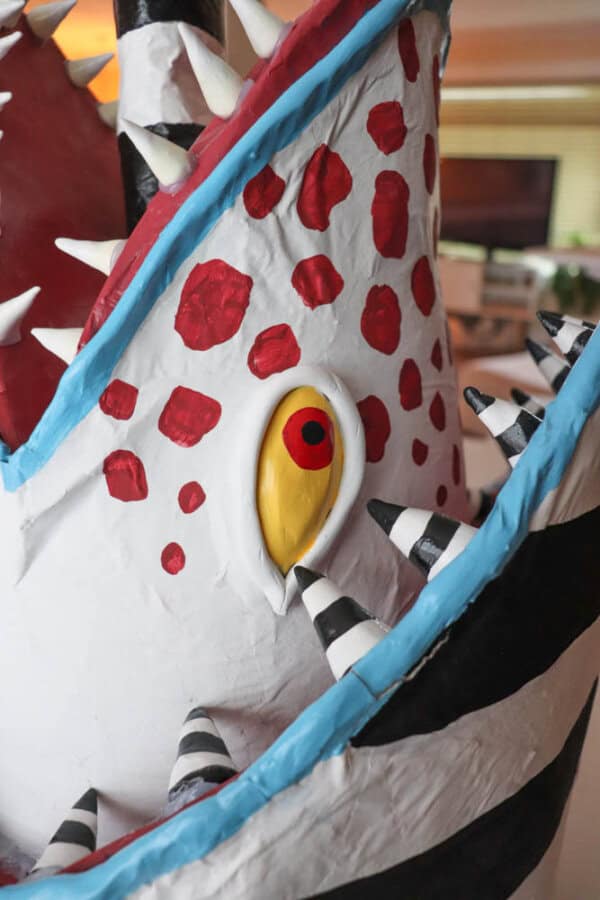 Close-up of a colorful, painted sculpture featuring red spots, a large yellow eye, and numerous spikes. The surface is white with black and white striped patterns along the edges. The background is blurred indoors.