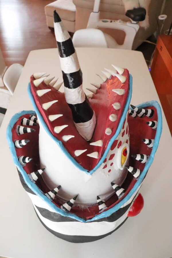 A colorful, whimsical mask resembling a sandworm with a striped horn, sharp teeth, and red mouth against a white background. The cakes body is striped in black and white, with eye-like patterns and a playful, artistic design.