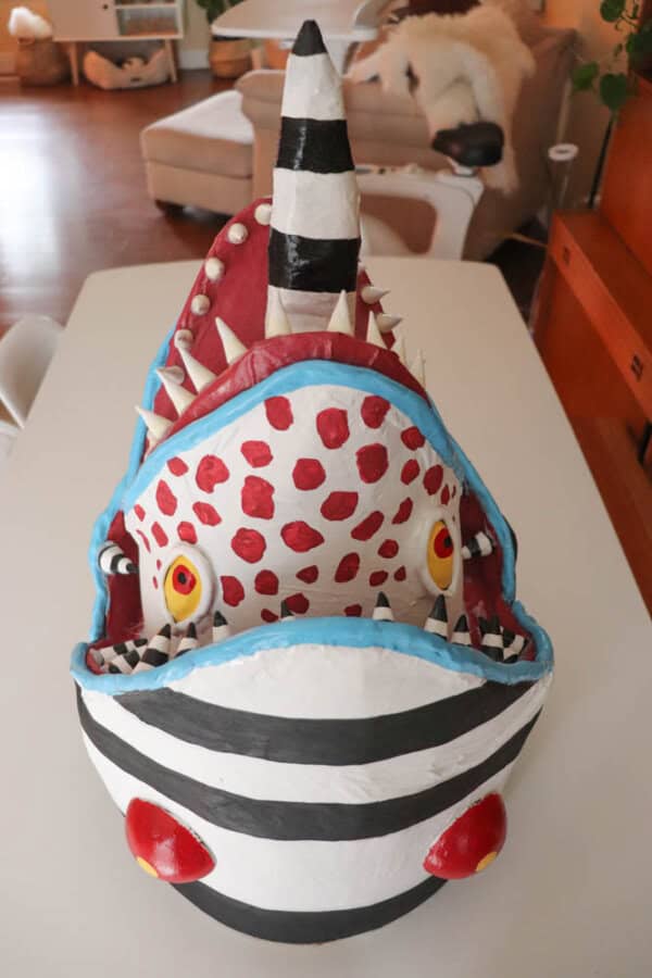 A colorful, handmade sculpture resembling a stylized shark with exaggerated features, including large red polka dots, sharp teeth, and big yellow eyes. The shark has a striped pattern on its body and sits on a table in a room.