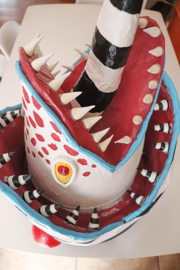 A sculpted mask designed to look like a sandworm with a large open mouth, sharp pointed teeth, red and white polka dots, and a striped black and white tongue sticking out.