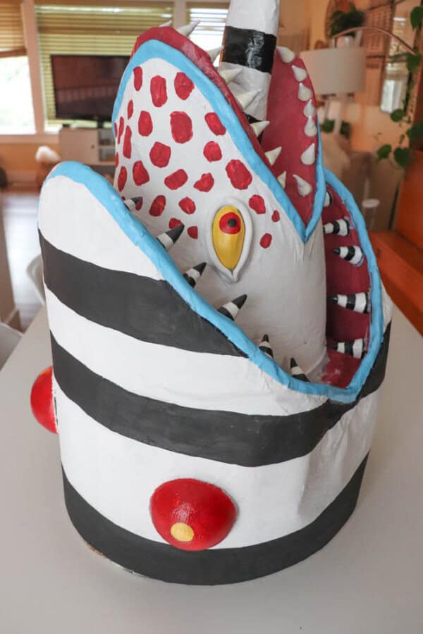 A colorful sculpture resembling a stylized, open-mouthed creature with red spots, yellow eyes, and striped black and white patterns. The sculpture stands on a white surface in a room with a window in the background.