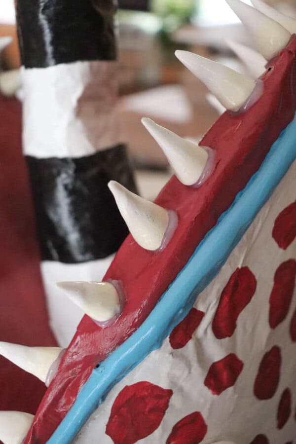 Close-up of a colorful paper mache sculpture featuring sharp white spikes along a red and blue edge. The background shows black and white striped and red patterned parts of the sculpture.