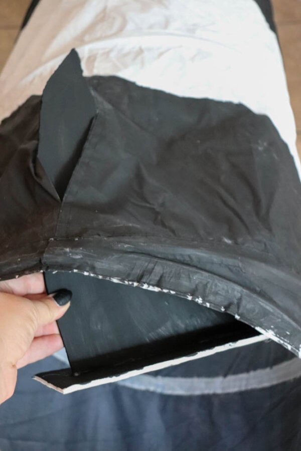 A person holding a slightly open flap of a black and white tunnel-like structure, showing its inner material. The structure appears to be made of fabric and is cylindrical in shape.