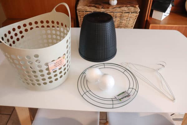 A white tabletop displaying a beige plastic basket, a black plastic container, two white foam balls on a circular wire frame, and two metal hangers. A wooden basket and shelves are visible in the background.
