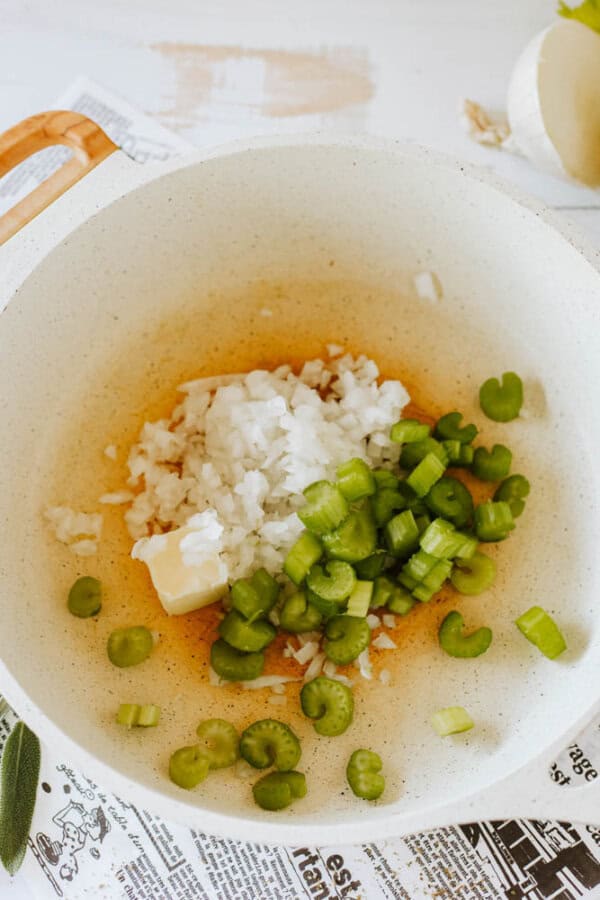 A large white pot contains chopped onions, celery slices, and a pat of butter. The ingredients are arranged on a brownish liquid, possibly oil or broth. A wooden handle is partially visible, and a newspaper is underneath the pot on a white surface.