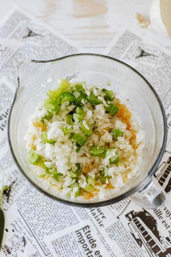 A glass bowl filled with chopped green peppers, onions, and other ingredients rests on a newspaper-covered surface. The mixture appears to be ready for cooking or mixing.