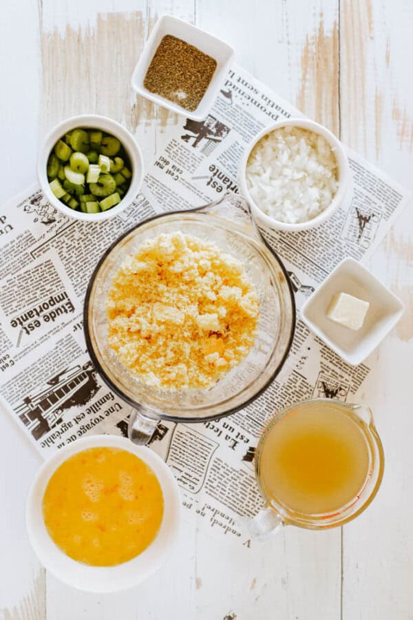 A flat lay of cooking ingredients on a newspaper. Includes a bowl of scrambled eggs, celery, onions, bread crumbs, pepper, butter, and a cup of liquid, likely broth, on a light wooden surface.