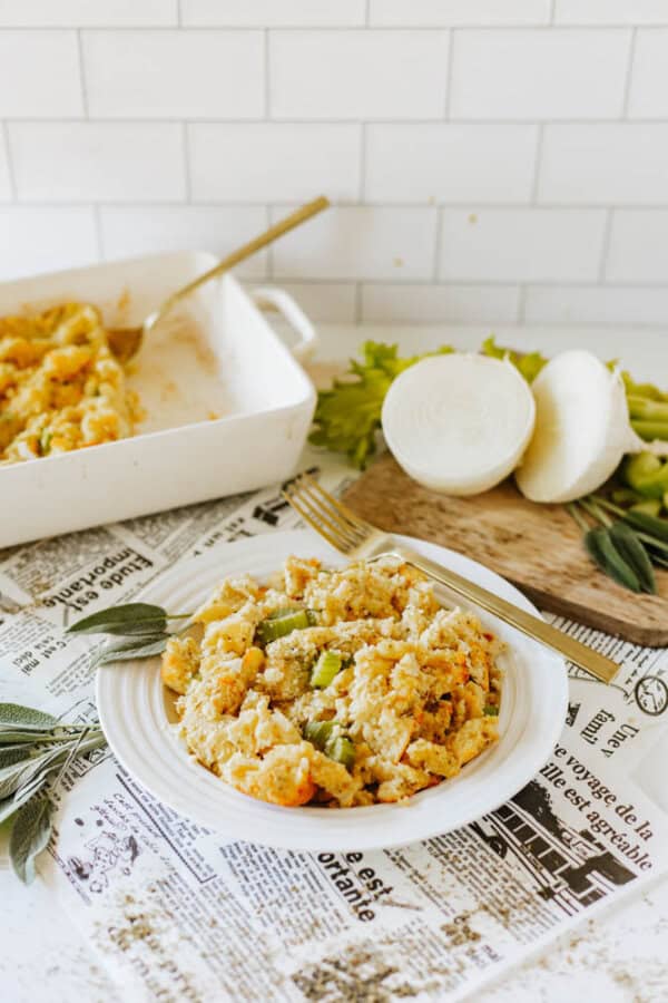 A plate of baked pasta with breadcrumbs and celery on a newspaper-lined surface. In the background, a baking dish with more pasta and a cutting board with sliced onion and celery are visible. Fresh sage leaves are placed nearby.