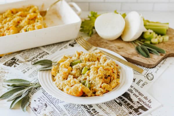 A plate of savory stuffing with celery and herbs is served on newspaper-themed paper. Behind it, a baking dish with more stuffing and a cutting board with sliced onion, celery, and sage leaves completes the scene. A fork rests on the plate.