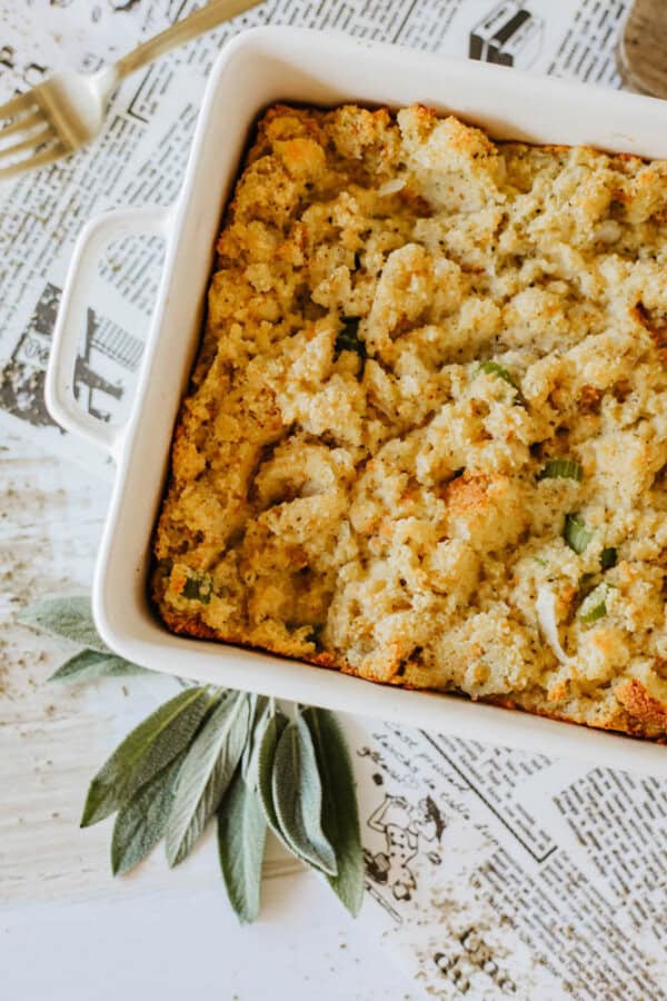 A casserole dish filled with golden-brown stuffing, topped with bread crumbs and herbs. Fresh sage leaves are placed beside the dish on a newspaper-covered table. A fork lies nearby.